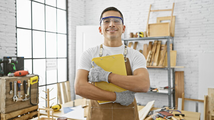 A confident young hispanic man in a carpentry workshop, holding a clipboard and wearing safety goggles and apron, represents professionalism and skill.
