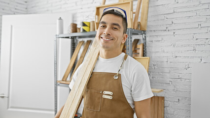 A smiling hispanic man in a carpentry workshop wearing protective goggles and a brown apron holding...