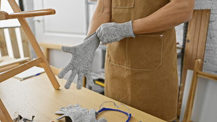 A hispanic man prepares to work by putting on gloves in a well-equipped carpentry workshop.