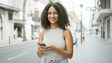 Joyful young hispanic woman brightly smiling while using her smartphone on a lively city street....
