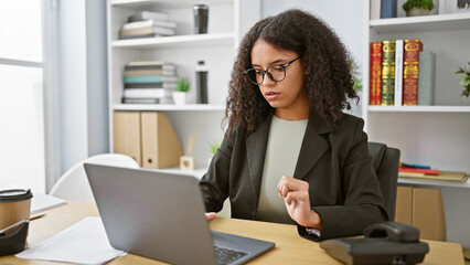 Striking curly-haired young hispanic woman, relaxed yet serious, elegantly working online on laptop...