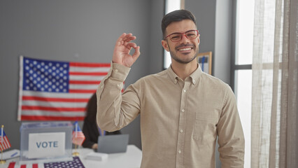 A smiling man gesturing ok with a woman and usa flag in a voting center backdrop.