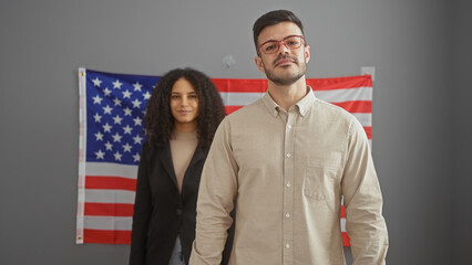A confident man and woman standing together indoors with an american flag in the background,...