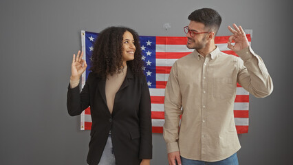 Man and woman gesturing 'ok' sign indoors with american flag backdrop symbolizing affirmation or...