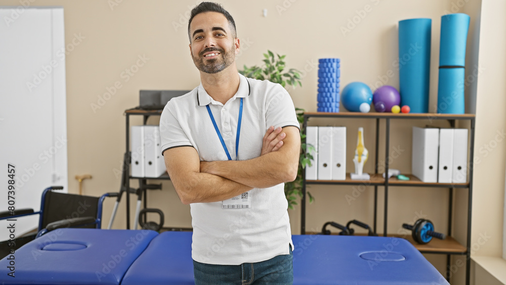 Canvas Prints Hispanic man with beard standing confidently in a well-equipped rehabilitation clinic, portraying professionalism and care.