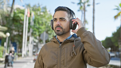 Handsome young hispanic man with beard wearing headphones in an urban city street setting.