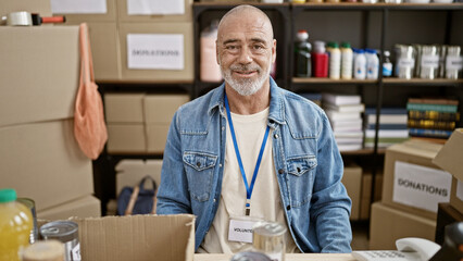 Smiling bearded man wearing volunteer badge stands in donation center surrounded by boxes and...