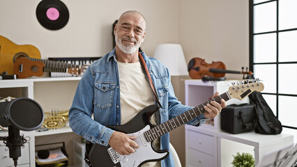 Mature man in denim playing electric guitar in a music studio with instrument background.