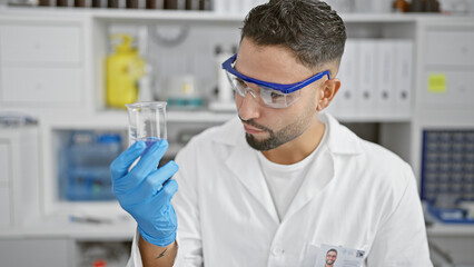 A focused man in lab coat and safety glasses examines a test tube in a science laboratory setting.