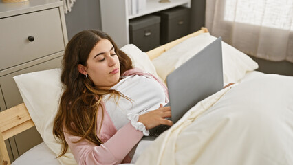 A young hispanic woman lounging in bed with a laptop, in a cozy bedroom setting, evokes relaxation...