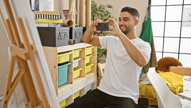 A handsome man capturing photo of his artwork with a smartphone in a creative indoor studio setting.