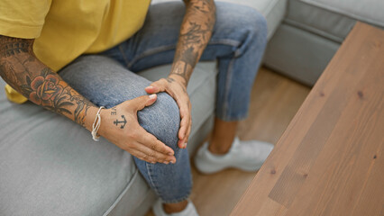 Hispanic man with tattoos sitting indoors grasping knee, potential pain at home.