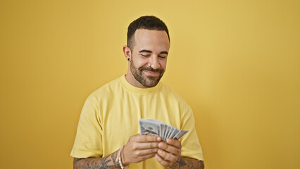 Handsome young hispanic man with a beard smiles as he counts money against a yellow isolated background.