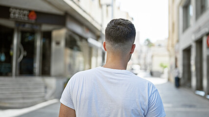 Fototapeta premium Back view of a young, hispanic man in casual clothing standing on an urban city street during daytime.