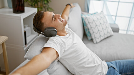 A relaxed young man wearing headphones reclining on a sofa with a content smile in a modern living room setting.
