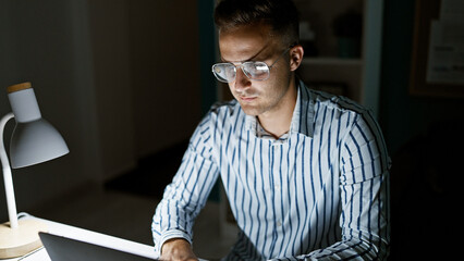 Handsome hispanic man working late on laptop in dark office room, exuding professionalism and focus.