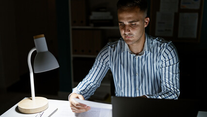 A focused hispanic man reading a document at night in a dimly lit modern office with a laptop and...