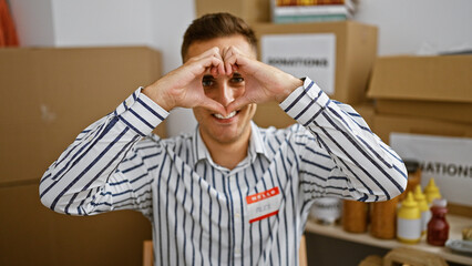 Young hispanic man making heart shape with hands in a warehouse setting, wearing striped shirt.