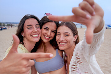 Happy three female friends on beach travel together, looking at camera smiling and gesturing a frame with fingers. Caucasian women enjoying summer in california.