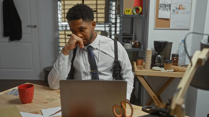 Pensive african american man in office, with laptop, coffee, board, badge, detective theme.