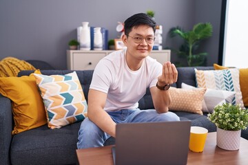 Young asian man using laptop at home sitting on the sofa doing money gesture with hands, asking for...
