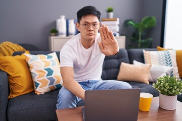 Young asian man using laptop at home sitting on the sofa doing stop sing with palm of the hand....