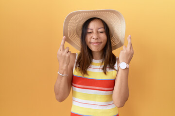 Middle age chinese woman wearing summer hat over yellow background gesturing finger crossed smiling...
