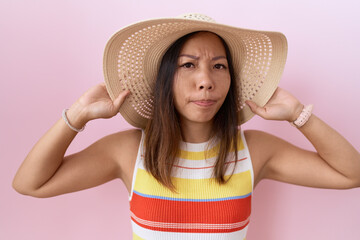 Middle age chinese woman wearing summer hat over pink background skeptic and nervous, frowning...