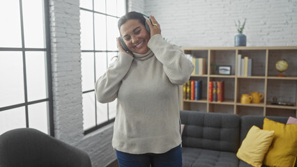 A joyful hispanic woman enjoys music on headphones in a cozy living room with bookshelf and sofa.