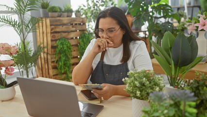 Middle-aged hispanic woman accounting in a green flower shop with laptop and lei banknotes.