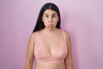 Young hispanic woman wearing pink bra puffing cheeks with funny face. mouth inflated with air, crazy expression.