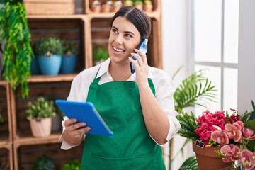 Young beautiful hispanic woman florist talking on smartphone using touchpad at flower shop