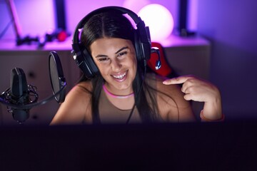 Young hispanic woman playing video games wearing headphones smiling happy pointing with hand and...