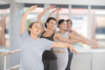 Group of women beginners rehearsing ballet moves at barre in dance studio