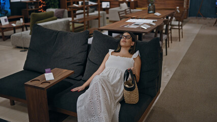 Beautiful hispanic woman in glasses choosing comfortable sofa at furniture store, comparing design...