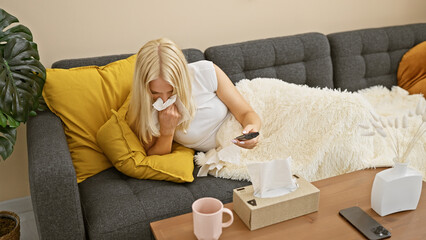 Stunning young blonde woman, ill with a nasty cold, lies beneath a cozy blanket on her homely sofa,...