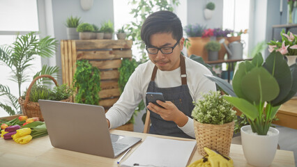 Young asian man using smartphone and laptop in a vibrant indoor flower shop surrounded by plants...