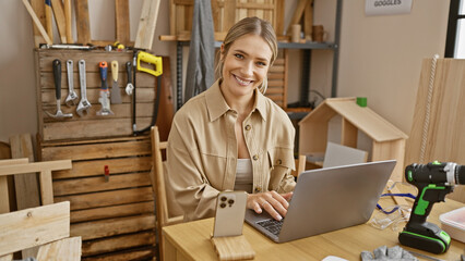 Attractive young blonde woman carpenter passionately immersed in online carpentry business on her laptop, smiling amidst the timbers in her cozy indoor workshop.