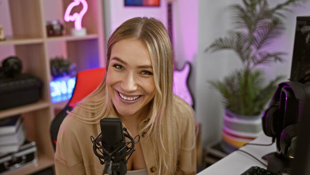 Smiling blonde streamer confidently speaking on a live gaming stream in her dark room, bringing virtual entertainment right to your house.