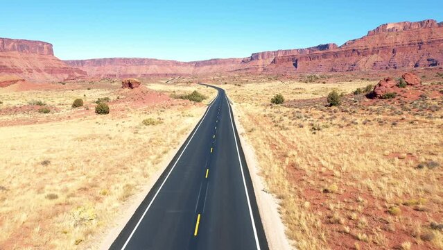 Empty intercity highway with black asphalt and yellow road markings pass through hot dry desert in western usa background of red rock massive butte in colorado river canyon. Aerial flying. Road trip