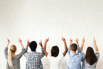 A group of friends stands in front of a blank white wall, all pointing upwards. Their gestures...
