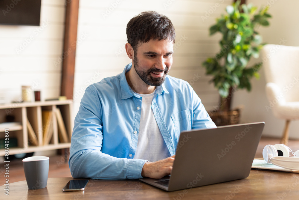 Aufkleber Businessman is seated at a table, focused on his laptop computer screen, typing on the keyboard as he works or browses online. The table is simple, with no distractions, and the background is plain.