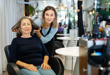 Professional hair stylist discussing elderly female customer preferences in salon, choosing new...