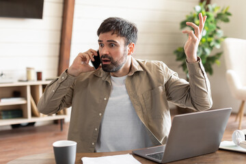 Man sits at wooden table, showing signs of frustration during phone conversation. His laptop is open, positioned in front of him, and he gestures with one hand, expressing his annoyance or confusion - obrazy, fototapety, plakaty