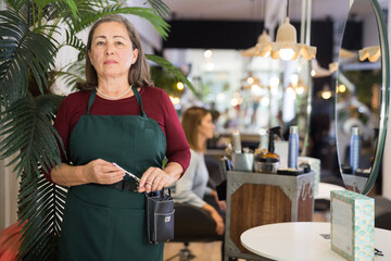 Portrait of experienced aged woman hairdresser wearing apron standing in modern barber shop with...