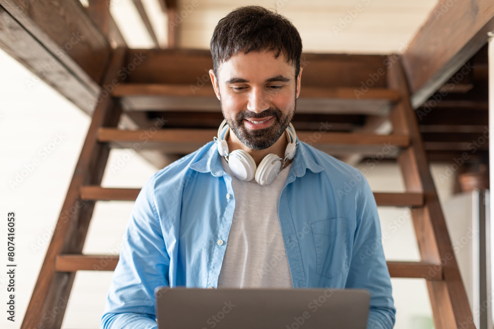 Poster a man is sitting on stairs, wearing headphones, and focused on using a laptop. he appears engaged in