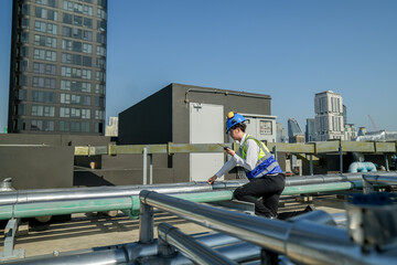 An engineer attentively checks pipework on a rooftop against a cityscape, using a tablet for data...