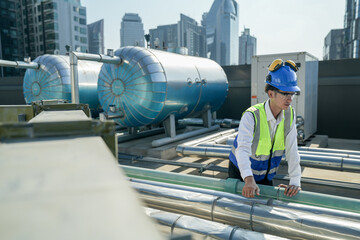 Naklejka premium Serious engineer inspecting HVAC equipment with a tablet on a commercial building rooftop. A focused engineer wearing a safety helmet and vest inspects machinery on a city building’s rooftop.