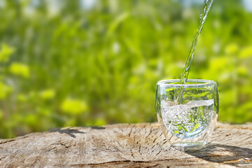 Water is poured into a glass against a background of greenery. A glass of water on a hot summer day...