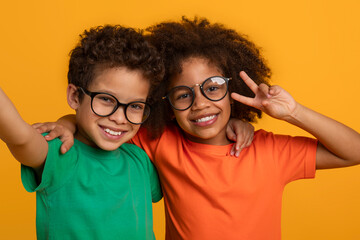 Two young African American kids, both wearing eyeglasses, are standing together and striking a pose for a photograph. They are smiling and looking directly at the camera